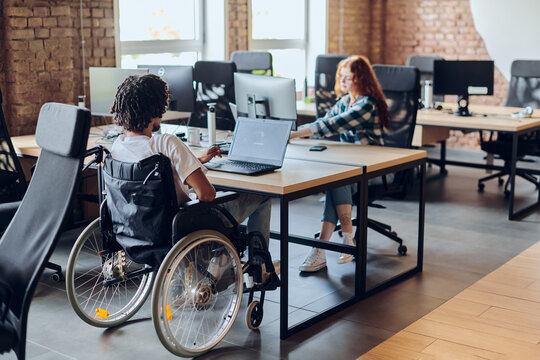 Business colleagues work within a modern startup coworking office, with one of them, an African America in a wheelchair, actively engaging with computers and laptops to collaboratively address various