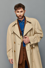 outerwear, trendy man in glasses and trench coat posing on grey backdrop, cozy layers, fall fashion