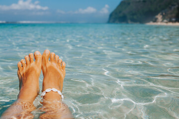 Tanned female legs with a white bracelet made of shells on a background of blue water. A stylish oceanfront holiday concept with space for text