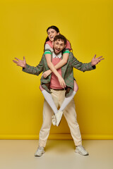 Obraz premium excited couple, funny, young man piggybacking brunette woman on yellow backdrop, student outfits