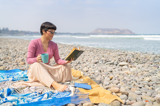 Woman on the beach reading a book and drinking coffee - Powered by Adobe