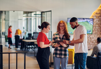 A group of young business individuals, including a girl with orange hair and an African American man, stands in a modern corporate hallway, collectively examining business progress on a smartphone