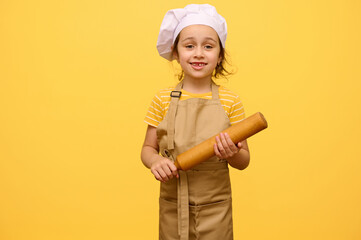 Happy little preschool girl with rolling pin, dressed as chef pastry, smiling looking at camera, yellow background