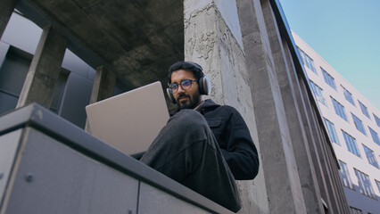 Indian guy university student in headphones listen business webinar or remote class studying outdoors Arabian man businessman male freelancer in city working online with laptop outside office building