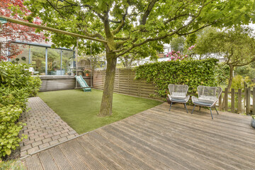 a backyard with wooden decking and green plants on the side of the house there is a tree in the yard
