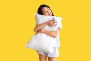 Beautiful Asian woman in pajamas with pillow on yellow background