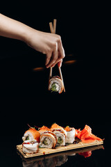 The Double Fish sushi roll in the wooden chopsticks in hand over the sushi with salmon, sesame next to which there is ginger, which are on a board made of wooden
