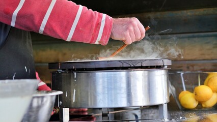 Cooking crepes at an outdoors event catering vendor, hand spreading pancake batter onto hotplate griddle pancake pan with a wooden crepe spreader, steam rising as the food cooks