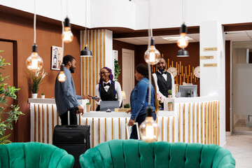 Young African American couple standing at hotel front desk with luggage, paying for room with...