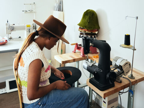  Black woman hat maker in a workshop space, sews on a sewing machine