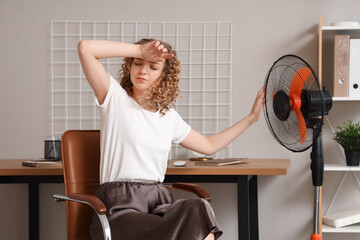 Beautiful young woman with electric fan sitting on chair at home during heatwave
