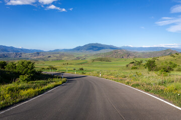 Winding tarmac road through Mtkvari river valley in Samtskhe - Javakheti region, Georgia with green fields around and Lesser Caucasus mountains in the background.