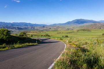 Winding tarmac road through Mtkvari river valley in Samtskhe - Javakheti region, Georgia with green fields around and Lesser Caucasus mountains in the background.