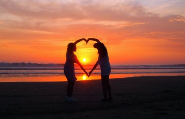 dos personas, haciendo un corazón con los brazos, a la orilla de la playa en un atardecer de...