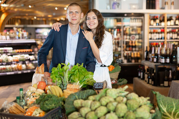 Glad couple is standing with cart with products in the supermarket