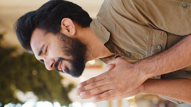 Bearded religion indian man praying with closed eyes to God asking for blessing, help, forgiveness outdoor. Adult guy clasping hands wishing luck in urban sunny city street. Town lifestyle. Vertical