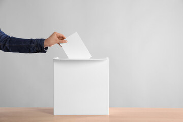 Woman putting her vote into ballot box on wooden table against light grey background, closeup. Space for text