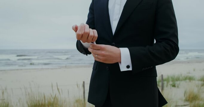 The groom fastens cufflinks on his shirt and smiles looking at the camera standing on the seashore