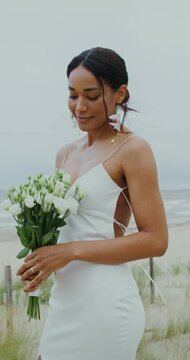 The bride smiles looking at the bouquet, and then at the camera, standing on the seashore