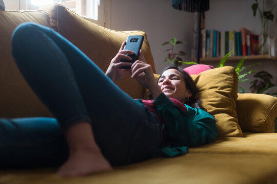 Happy lazy woman using mobile phone lying on the sofa