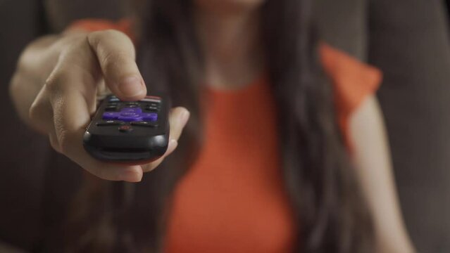 Unrecognizable woman using the remote control to change the channel. Sitting on the couch on an orange dress. SHOTLISTstream.