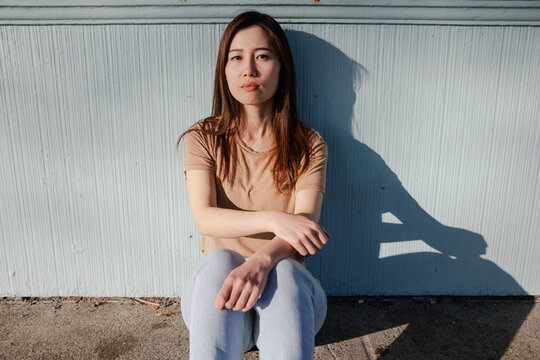 Young woman in casual clothing sitting against a blue wall.