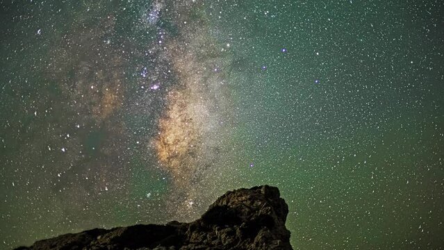 Timelapse Depicts the Milky Way Galaxy's Movement Across the Night Sky, Infusing Rocks Below with a Soft Emerald Radiance.