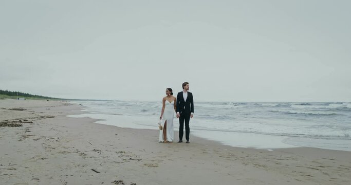 The bride and groom hold hands standing on the seashore and look in different directions