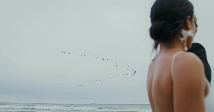 The bride and groom are walking along the seashore, a flock of birds is flying across the sky