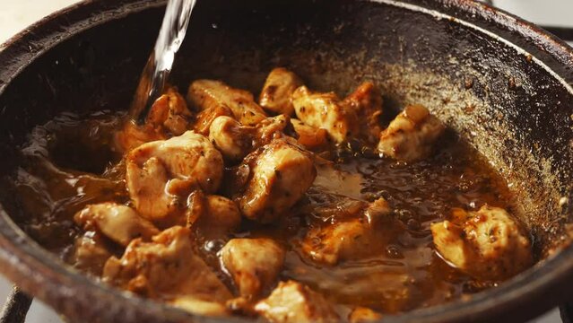 Chicken pieces fried in a cast-iron skillet in oil with onions and spices are poured with water. Close-up
