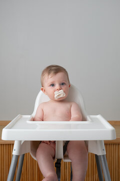 Baby with white pacifier sits in white highchair.