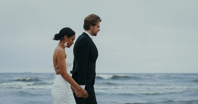 The bride and groom hold hands and smile at each other while walking on the beach