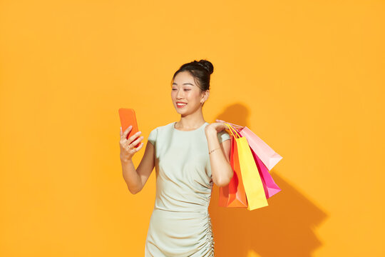 happy young woman holding shopping bags and mobile phone over yellow