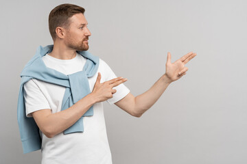 Positive young caucasian man looking and pointing fingers aside, gesturing at copy space on product, sale discount promo offer standing isolated on grey background. Advertising banner, poster concept.