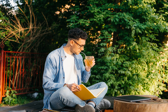 A man drinks a soda sitting outdoors with a notebook in his hands