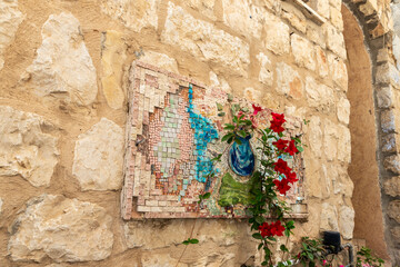 A large  decorative mosaic on the stone wall of a residential building and flowers standing nearby on a quiet street in the old part of Safed city in northern Israel