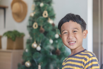 Close-up Asian boy with black hair wearing a yellow-gray striped t-shirt looking at the camera in the background of christmas trees and wooden furniture. Child makes funny faces in his house happily.