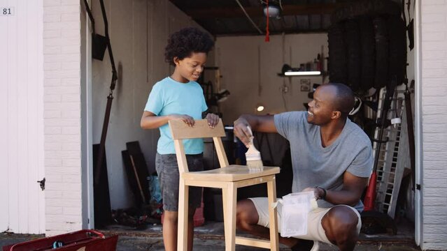 Mixed race father teaching son how to varnish the wooden chair at home during DIY project in the garage
