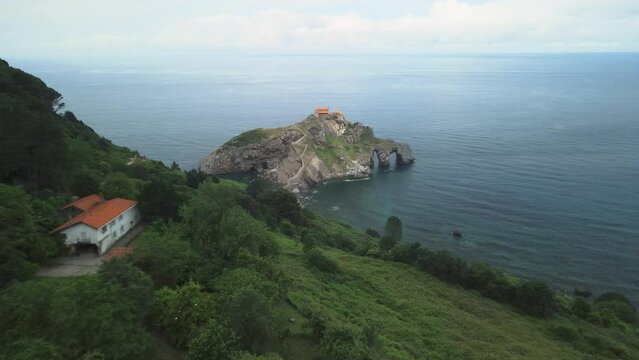 Drone descends along lush green sea cliffs to monastery on Gaztelugatxe Basque Spain, establishing