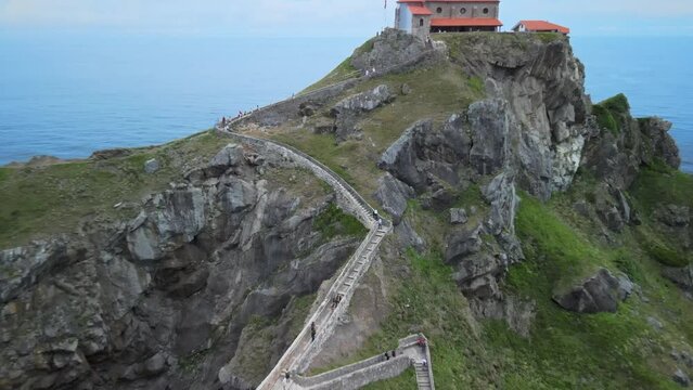Drone pullback reveals Monastery on Gaztelugatxe Island, green sea cliffs and calm ocean waves, Basque Spain