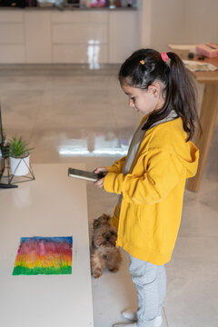 Girl Captures Watercolor Drawing with Phone, Dog Curiously Watches.