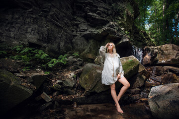 A smiling happy pregnant girl in a light vintage dress, with blond short hair, stands against the backdrop of picturesque rocks near a waterfall in the forest.