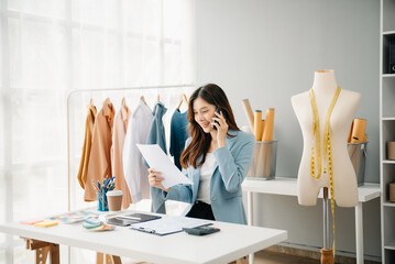 Fashion designer woman talking smart phone and using laptop with digital tablet computer in modern studio the clothes hanging on the racks