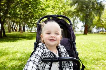 Baby in stroller on a walk in summer park. Adorable little girl sitting in pushchair and smiles. Child in buggy