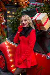 A girl in a red coat and beret on the background of a car with gifts and a Christmas tree with lights
