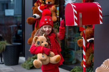 A child girl dressed in red stands next to a mailbox and holds a teddy bear in her hands. Merry Christmas - Powered by Adobe