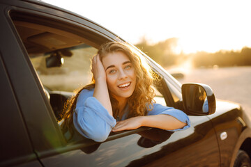 Smiling woman from the window of the car enjoys nature, you feel freedom while traveling by car. Car travel, sunset light. Active lifestyle.