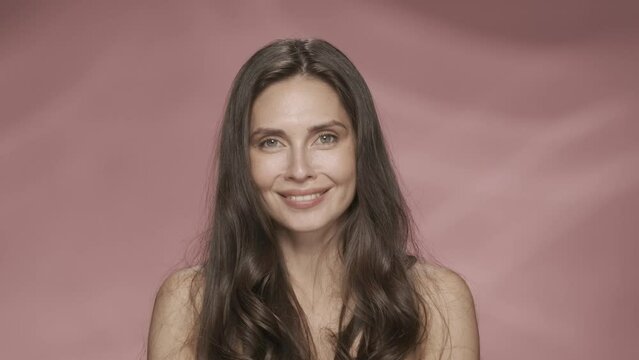 Portrait of a seminude brunette woman with flowing hair in the studio on a pink background. The woman holds up her palms with yellow rose petals and blows them away. HDR BT2020 HLG Material.