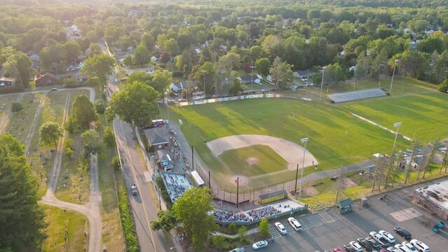 A baseball aerial shot heading away from the field. The field is outside surrounded by trees with bleaches near the field and a road near by with cars driving by.