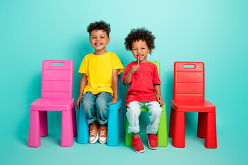 Full body photo of cute little boys eating sweet lollipops sit chairs wear trendy clothes isolated...